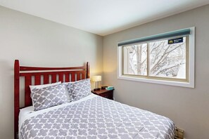 Ein Schlafzimmer mit einem hölzernen Bett, gemusterten Betttextilien, einem Nachttisch mit Lampe und einem Fenster mit Blick auf Bäume.