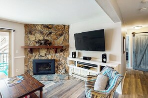 Ein Wohnzimmer mit Kamin aus Stein, einem Flachbildfernseher, einem Couchtisch aus Holz und einem gestreiften Sessel.