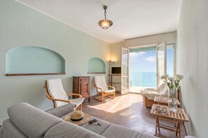 Ein Zimmer mit einer Couch, zwei Schaukelstühlen, einem kleinen Tisch mit einer Vase und Blumen sowie Blick auf das Meer durch eine offene Tür.