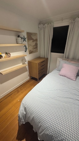 A bedroom with a bed, wooden dresser, shelves, and a window with curtains.. Bach over the bay