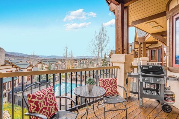 Eine Holzterrasse mit Sitzgelegenheit, Grill und Blick auf einen Swimmingpool sowie Gebäude im Hintergrund.