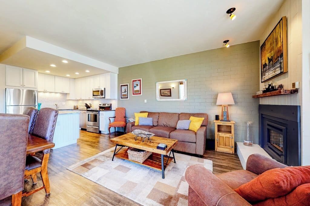 A modern living room with a sofa, coffee table, and chairs, a kitchen in the background, and a fireplace.