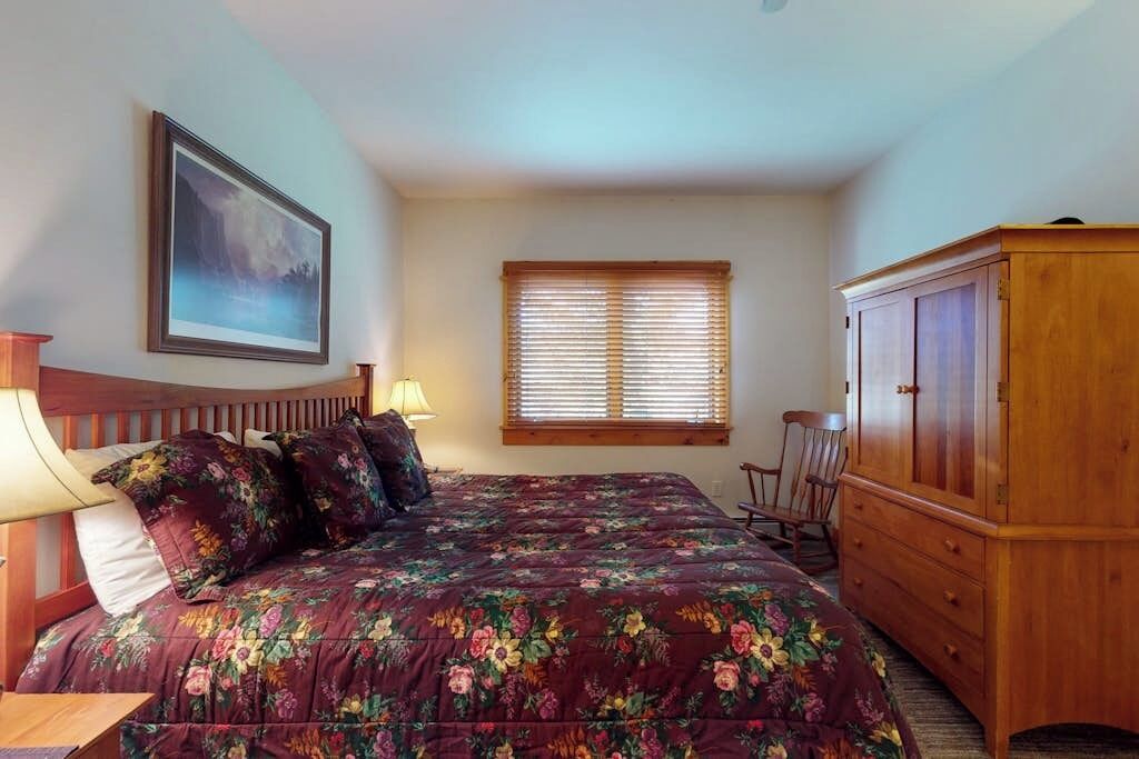 Un dormitorio con una cama, un armario de madera, una ventana con persianas, una silla y un cuadro en la pared.