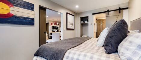 A bedroom with a bed, a wall-mounted artwork, a door leading to a bathroom, and a hallway.