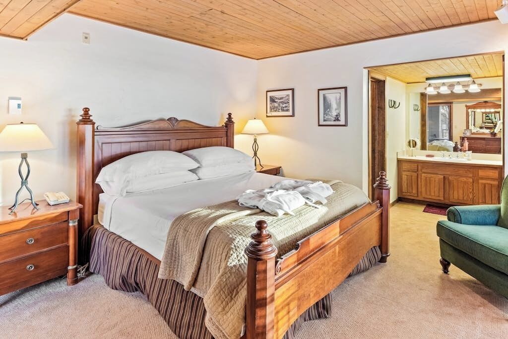 A bedroom with a wooden bed, bedside lamps, a green armchair, and a bathroom with a mirror and vanity.
