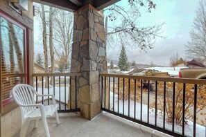 A balcony with a stone pillar, a white plastic chair, and a view of a snowy landscape with trees and a parked car. - Walk-to-gondola condo w/ fireplace, W/D & resort pool/hot tubs/shuttle/gym (Steamboat Springs)