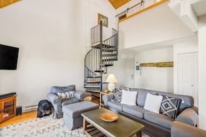 Ein Wohnzimmer mit grauem Sofa, einem kleinen Tisch, einer schwarzen Wendeltreppe und einem an der Wand befestigten Fernseher.