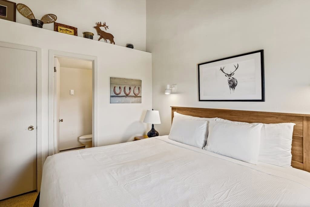 A hotel room with a large bed, a wooden headboard, a framed deer head picture, a wall-mounted lamp, and a door leading to a bathroom.