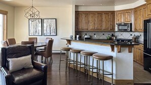 A modern kitchen with wooden cabinets, a central island with stools, a dining area with leather chairs, and a hanging light fixture. - Walk-to-lift townhome w/ fireplace, laundry & resort pool/hot tubs/gym/firepit (Steamboat Springs)