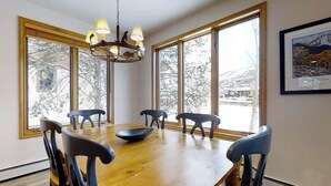 A dining room with a wooden table, six chairs, a chandelier, large windows, and a framed picture on the wall. - Evergreen 03 Luxurious Townhome Featuring a Shared Pool and Shuttle (Steamboat Springs)