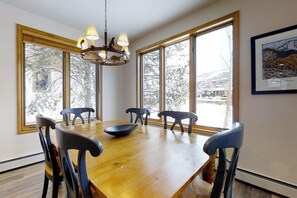 A dining room with a wooden table, six chairs, a chandelier, large windows, and a framed picture on the wall. - Luxurious Townhome Featuring a Shared Pool and Shuttle (Steamboat Springs)