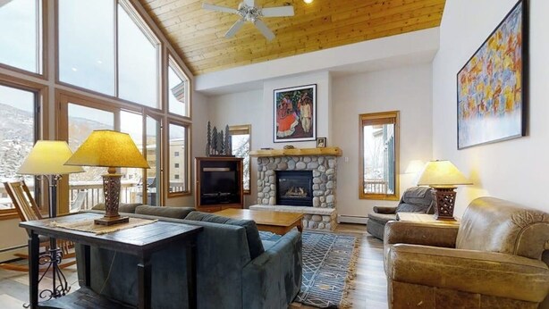 A spacious living room with a stone fireplace, large windows, and a ceiling fan. - Evergreen 03 Luxurious Townhome Featuring a Shared Pool and Shuttle (Steamboat Springs)