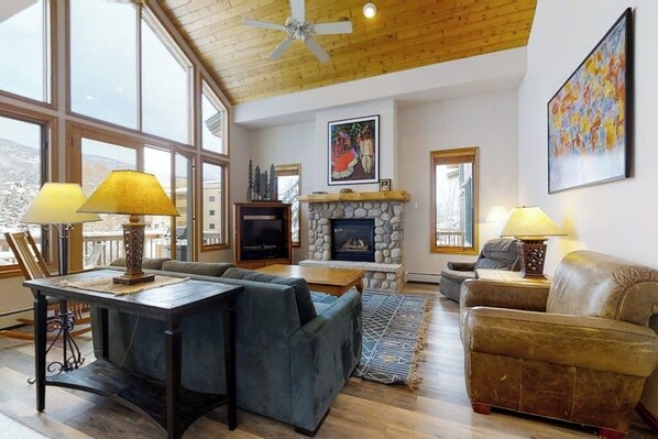 A spacious living room with a stone fireplace, large windows, and a ceiling fan. - Luxurious Townhome Featuring a Shared Pool and Shuttle (Steamboat Springs)