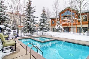 Ein Schwimmbecken mit Holzterrasse, umgeben von schneebedeckten Bäumen und einem Gebäude mit Balkon.