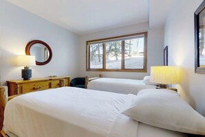 Ein Hotelzimmer mit zwei Betten, einem hölzernen Schminktisch, einem runden Spiegel, einem Fenster mit Blick auf Bäume und einer Lampe.