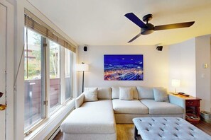 Ein modernes Wohnzimmer mit Ecksofa, Deckenventilator, großem Fenster mit Jalousien und Stadtbildgemälde an der Wand.