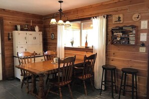 A wooden interior with a dining table, chairs, a bar stool, a refrigerator, and a window with curtains.