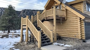 Une cabane en rondins avec un escalier en bois menant à un balcon, le tout entouré d’un paysage enneigé.