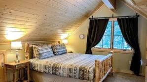 Une chambre avec un plafond en bois, un lit avec une literie à motifs, une table de chevet, une lampe et une fenêtre avec des rideaux.