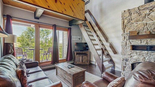 A spacious living area with a stone fireplace, a wooden staircase, and a large sliding glass door leading to an outdoor deck.