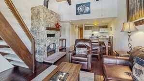 A spacious living area with a stone fireplace, leather furniture, a staircase, and a kitchen in the background.