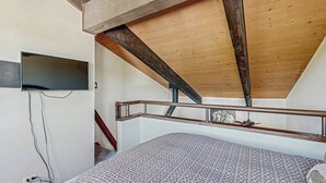A bedroom with a sloped ceiling, a bed, a television mounted on the wall, and a staircase.