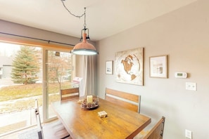 A dining area with a wooden table, chairs, a hanging pendant light, and a window with a view of trees and buildings. - Mountain condo with full kitchen, fireplace, & deck - close to skiing (Steamboat Springs)