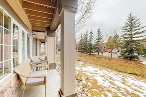 A covered outdoor seating area with wicker furniture, a stone wall, and a view of a snowy landscape with trees. - Mountain condo with full kitchen, fireplace, & deck - close to skiing (Steamboat Springs)