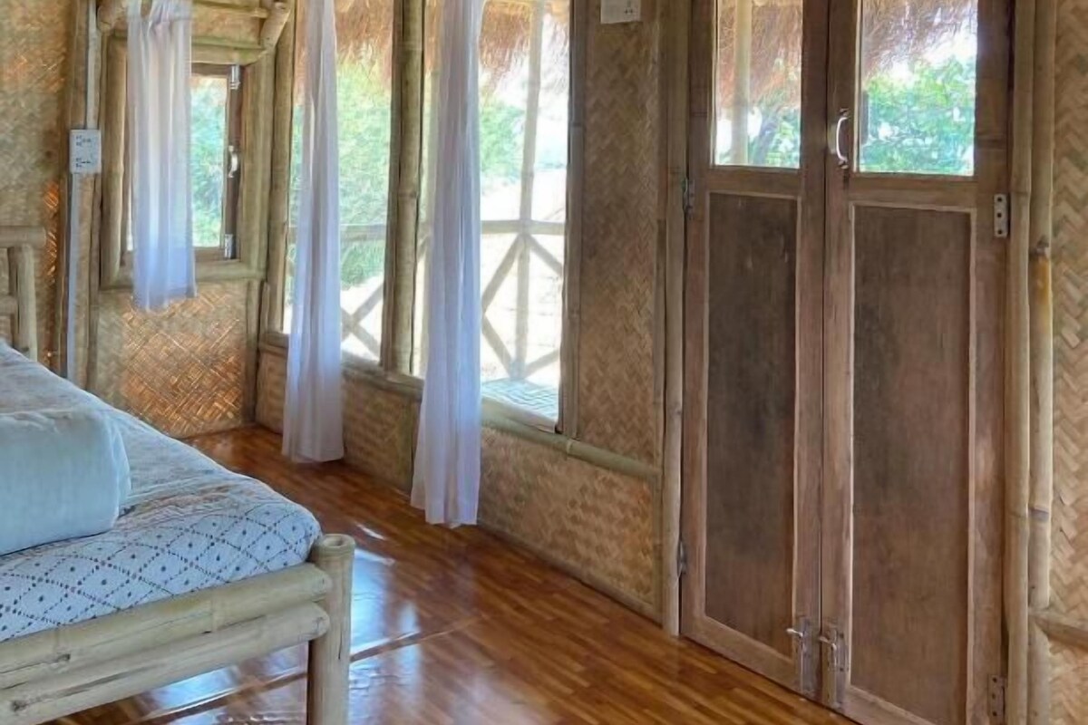 Una habitación con cama, piso de madera, ventanas grandes con cortinas y puerta de madera.