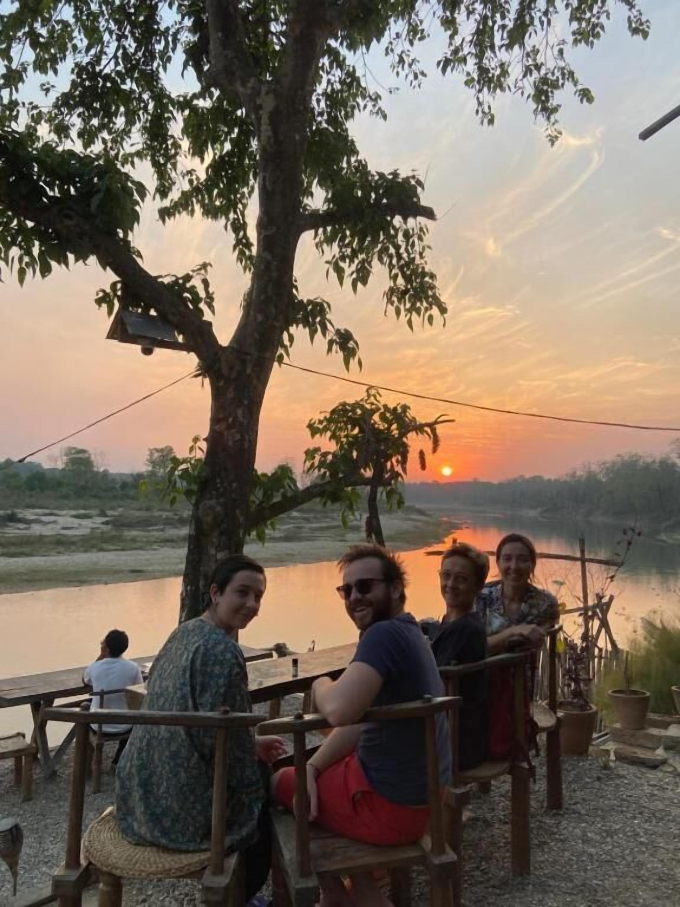 A group of people seated at wooden tables outdoors with a sunset in the background.