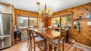 Un intérieur en bois comprenant une table à manger, des chaises, un lustre, un réfrigérateur et de grandes fenêtres.