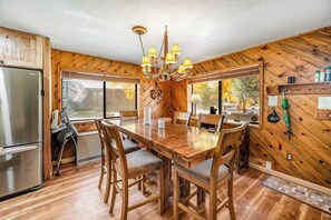 Ein Holzinterieur mit einem Esstisch, Stühlen, einer Chandelieber, einem Kühlschrank und großen Fenstern.