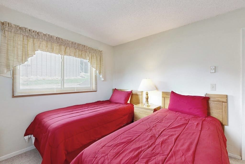A room with two beds, a nightstand with a lamp, and a window with blinds.