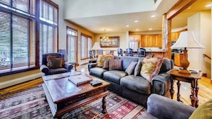 A spacious living room with a large sectional sofa, a coffee table, and a rug. There are large windows with blinds, a kitchen in the background, and a chandelier. - 1442 EagleRidge Walk to the gondola Townhome w/ deck, fireplace, W/D & shared hot tubs/pool (Steamboat Springs)