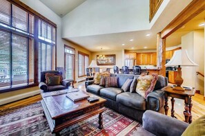 A spacious living room with a large sectional sofa, a coffee table, and a rug. There are large windows with blinds, a kitchen in the background, and a chandelier. - Walk to the gondola Townhome w/ deck, fireplace, W/D & shared hot tubs/pool (Steamboat Springs)
