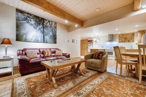 Ein Wohnzimmer mit einem roten Ledersofa, einem beigefarbenen Sofa, einem Holztisch und einem Teppich. An der Wand hängt ein Bild von einem Wald, und im Hintergrund ist eine Küche zu sehen.