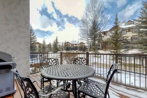 Ein Balkon mit einem runden Metalltisch und Stühlen, einem Grill und Blick auf schneebedeckte Bäume und Gebäude.