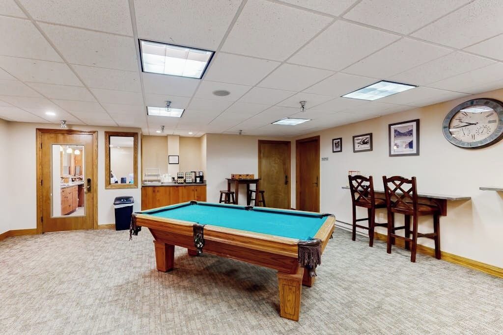 A pool table in a room with a carpeted floor, wooden chairs, a clock on the wall, and framed pictures.