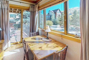 Ein Zimmer mit Holztisch und Stühlen, großen Fenstern mit Vorhängen und Blick auf Gebäude und Bäume.