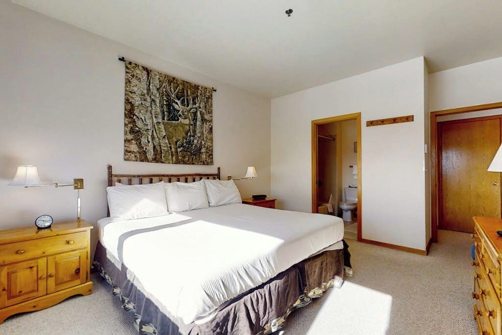 A bedroom with a large bed, a nightstand, a wall-mounted painting, a wooden wardrobe, and a door leading to another room.