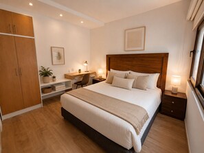 A bedroom with a large bed, a desk, a chair, a wardrobe, and a bedside table with a lamp.