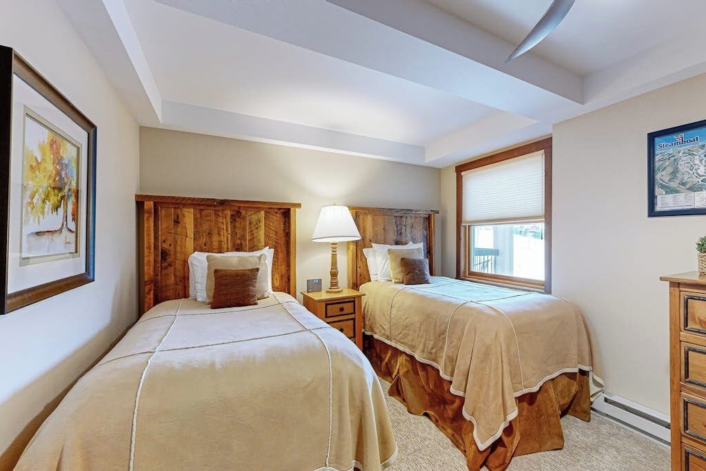 Une chambre d’hôtel avec deux lits, des têtes de lit en bois, une table de chevet avec une lampe, une fenêtre avec des stores et une carte encadrée accrochée au mur.