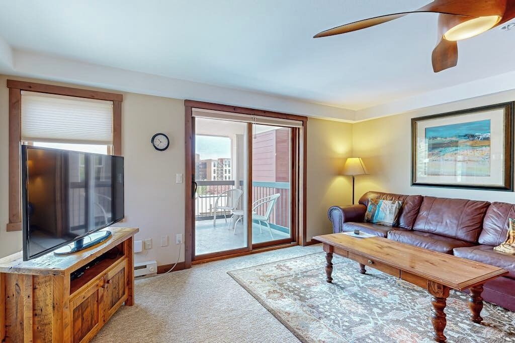 A living room with a flat-screen TV on a wooden stand, a leather sofa, a wooden coffee table, a patterned rug, a balcony with a view, and a painting on the wall.