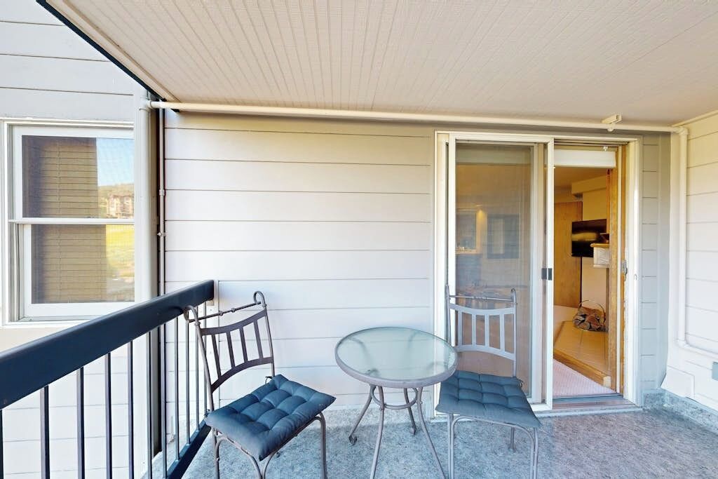 A balcony with a glass table and two chairs, a sliding glass door leading to an interior room, and a white exterior wall.