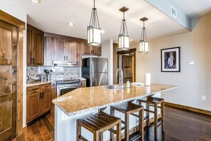 A modern kitchen with wooden cabinets, stainless steel appliances, a granite countertop, and bar stools. - Brilliant upscale condo w/ADA accessibility & shared hot tubs, pool, gym, grills (Steamboat Springs)