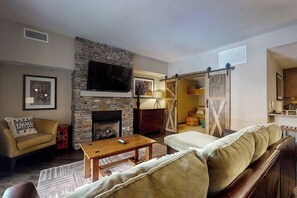 A living room with a stone fireplace, a sofa, a coffee table, and a TV mounted above the fireplace. - Beautiful condo w/bonus den & shared pool, hot tubs, game room, fitness & more (Steamboat Springs)