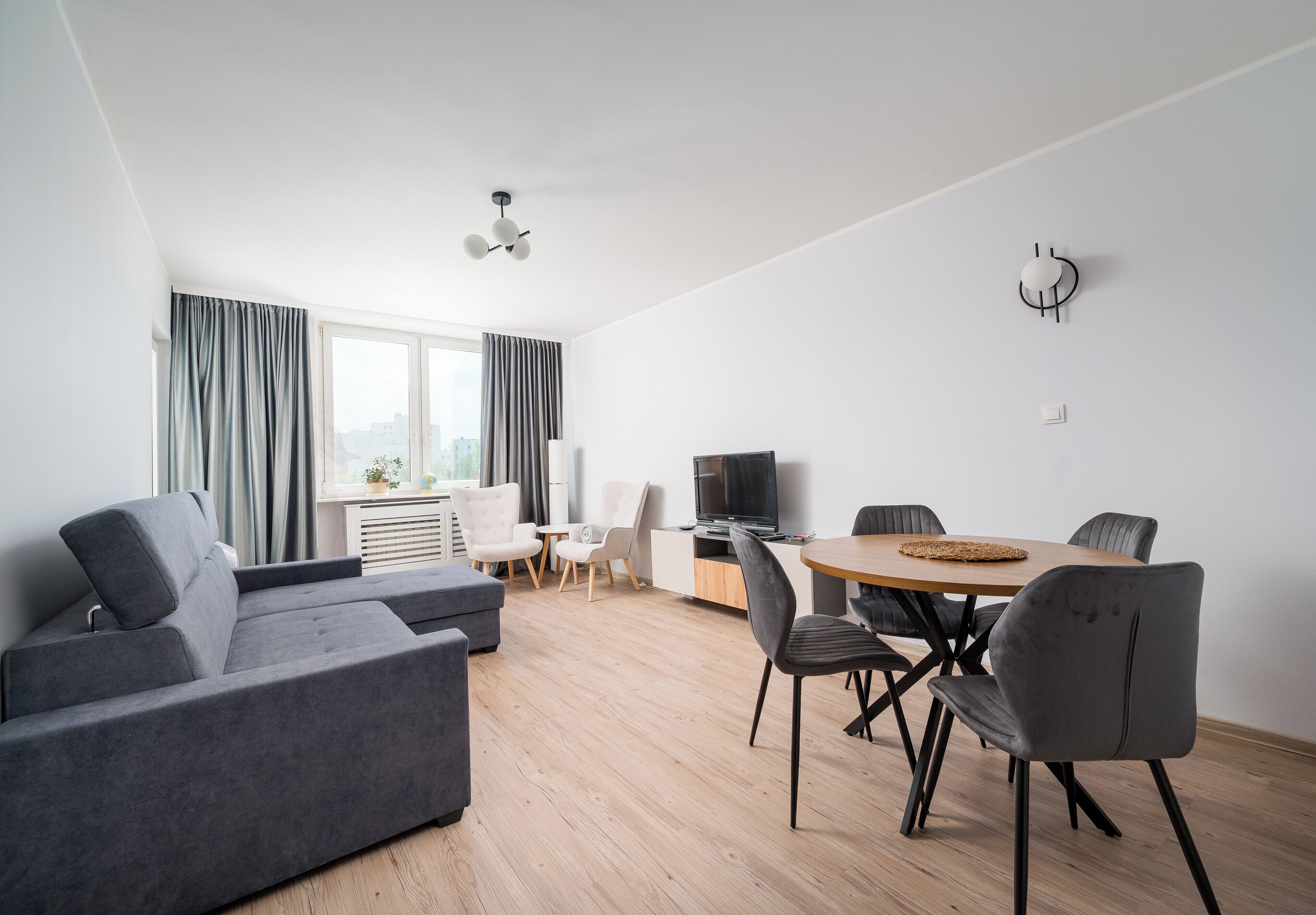 Ein modernes Wohnzimmer mit einer Couch, einem Esstisch, Stühlen, einem Fernseher und einem Fenster mit Vorhängen.