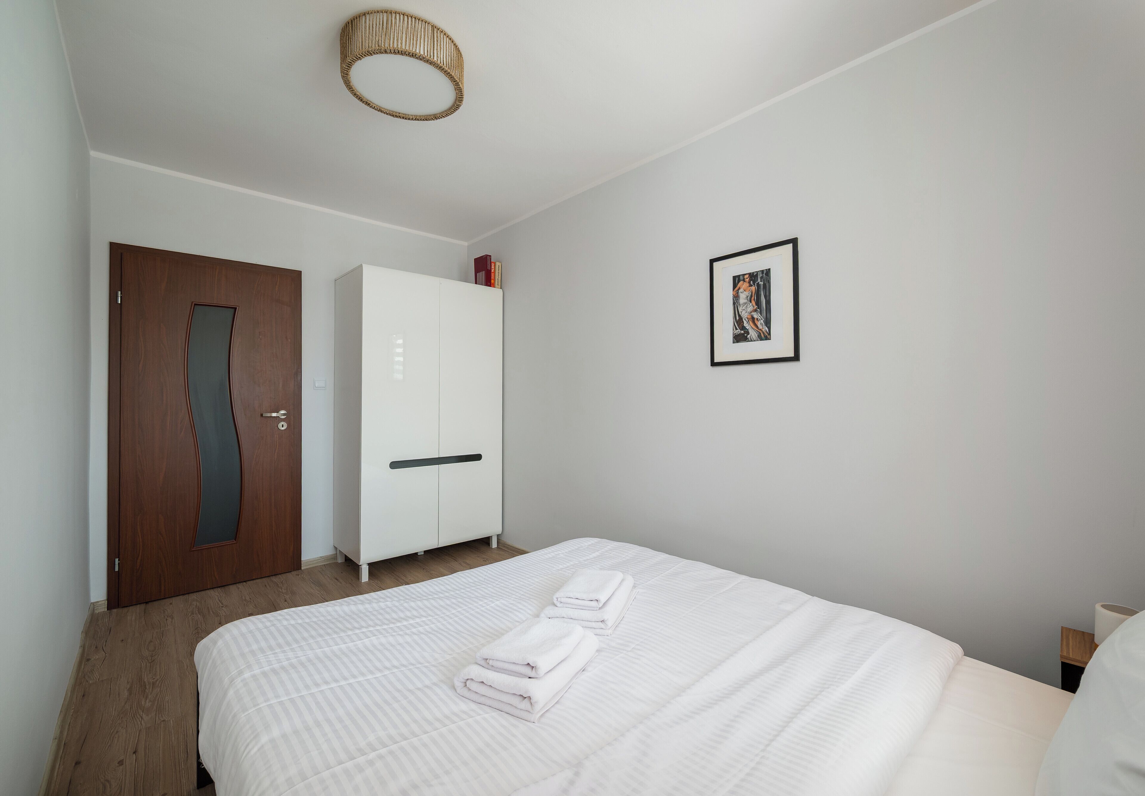 Una camera da letto con un letto, tende bianche, una porta in legno, un armadio e un quadro appeso al muro.