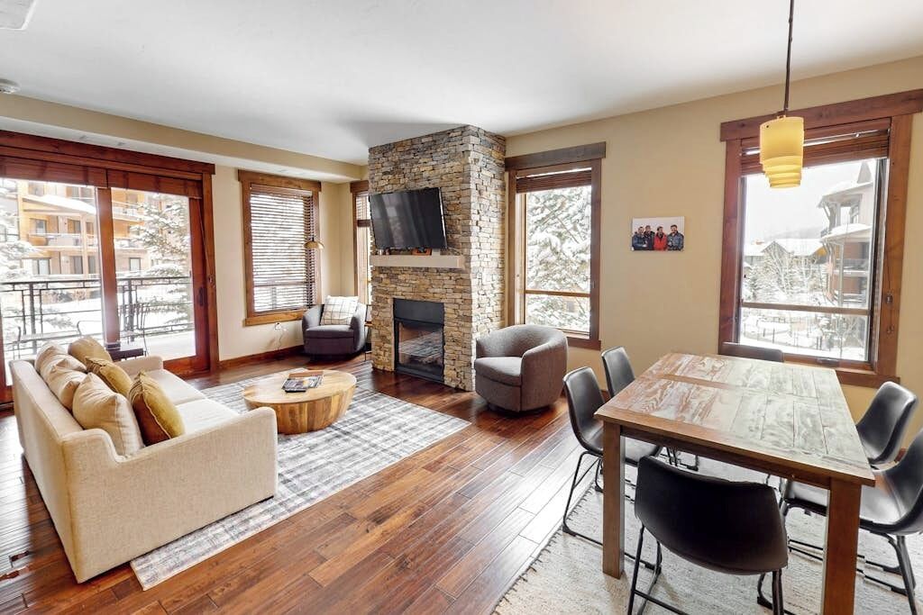 A living room with a stone fireplace, a sofa, a dining table with chairs, and large windows.