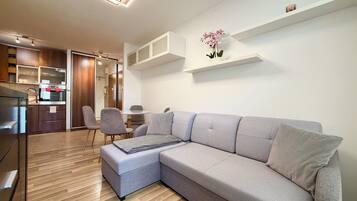 A modern living room with a grey sectional sofa, a small round table, and chairs. There is a kitchen area with wooden cabinets and a built-in oven. A potted orchid is on a shelf above the sofa.